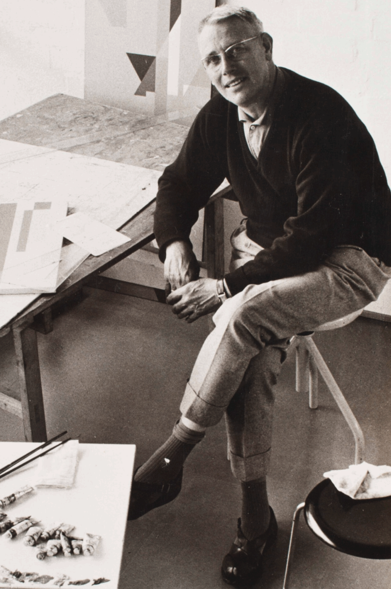 A man seated on a chair in a studio, wearing a black sweater and glasses, with one leg crossed over the other. He is surrounded by art materials on a table, including paint tubes and brushes.