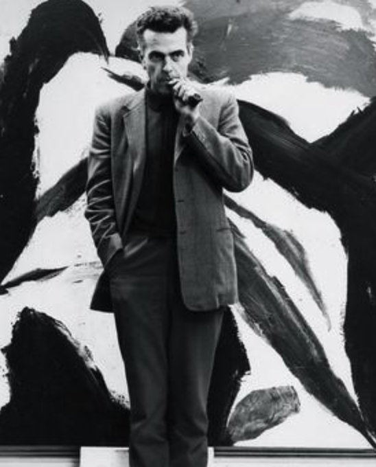 A man in a suit stands against a large black and white abstract painting, holding a cigarette to his mouth.
