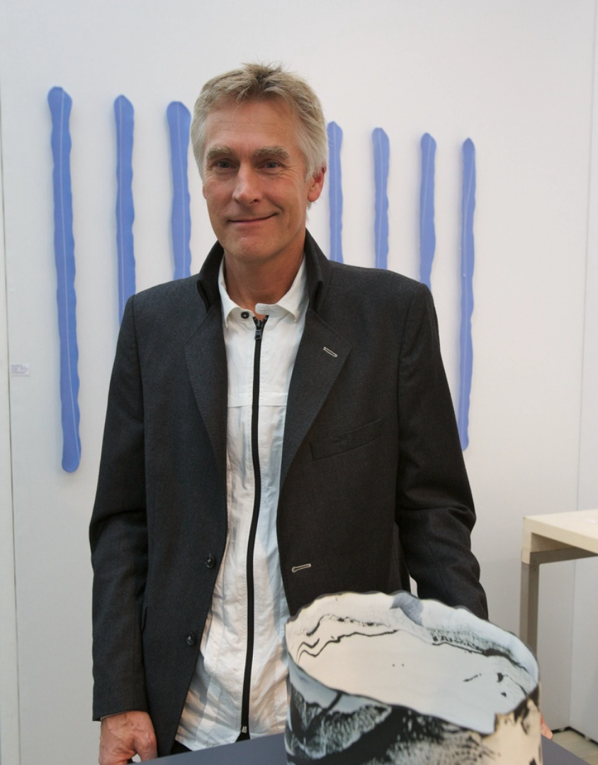 A smiling individual wearing a dark jacket stands in an art gallery next to a unique ceramic sculpture, with blue vertical installations in the background.