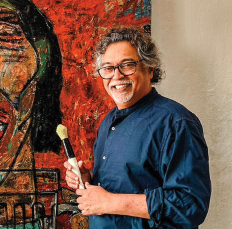 An artist smiling while holding a paintbrush, standing in front of a vibrant, colorful painting.