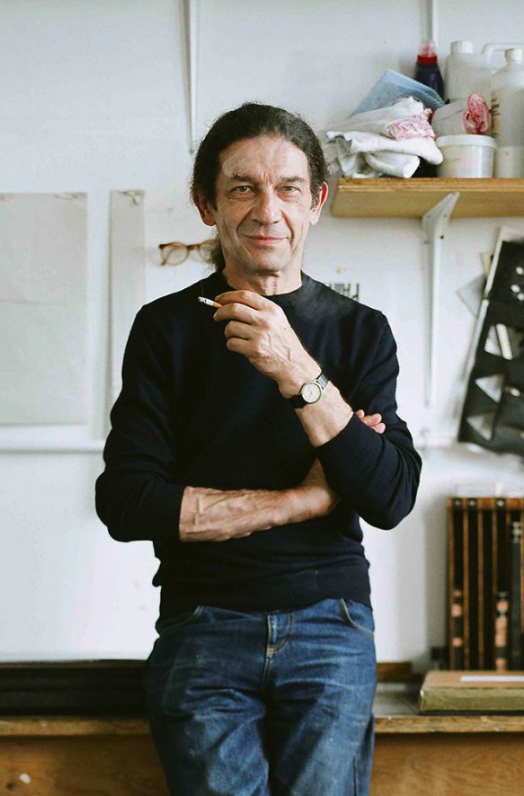 A man with long hair stands against a workbench, arms crossed and holding a cigarette, wearing a black sweater and jeans, with art supplies and papers visible in the background.