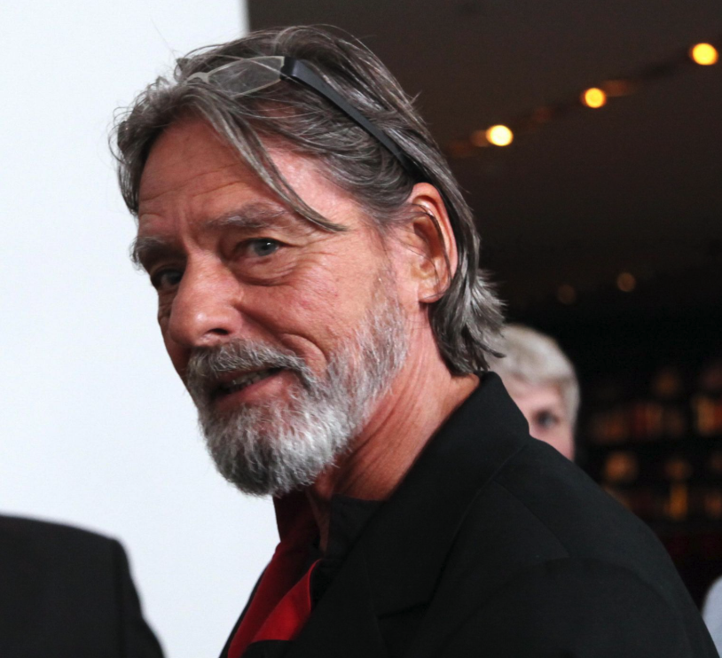 Close-up of a man with long, gray hair and a beard, wearing glasses on his head and a black jacket, smiling slightly in an indoor setting.