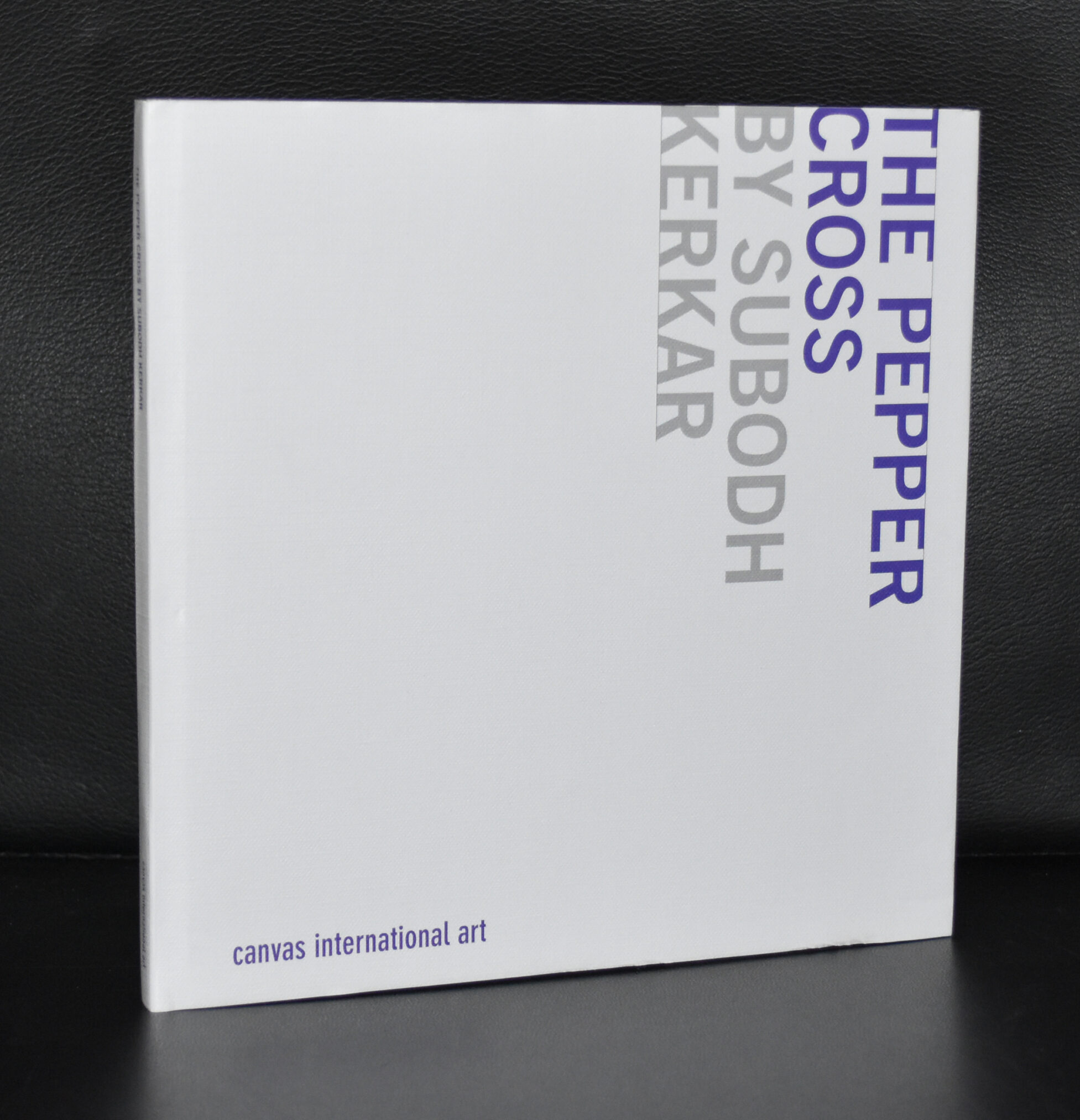 A book titled 'The Pepper Cross by Subodh Kerkar', with a minimalist white cover displaying the title in bold gray and purple letters, resting on a black surface.