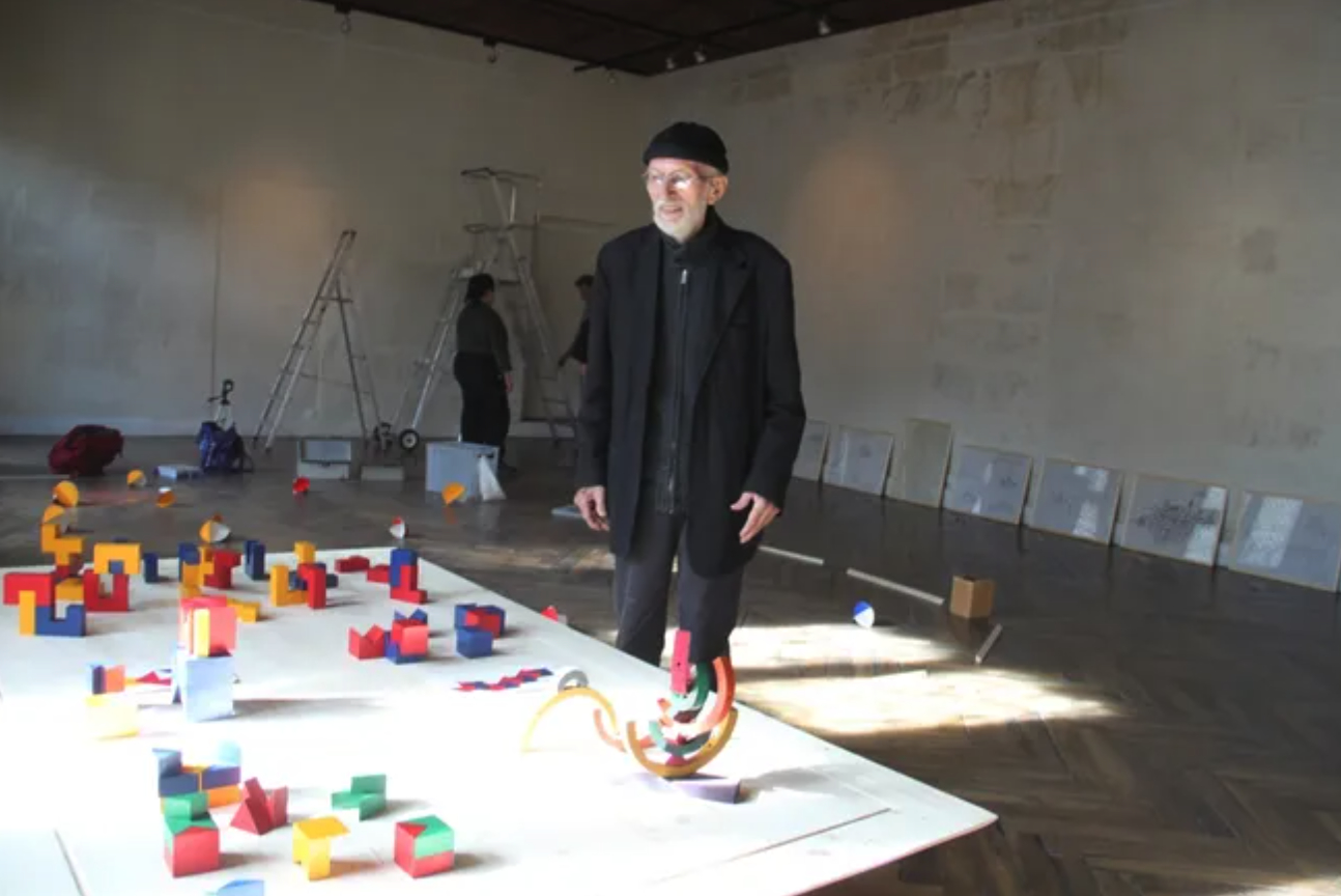 Claude Pasquer standing in an art studio with colorful geometric shapes scattered on a table and around him, engaging with the space and artwork.