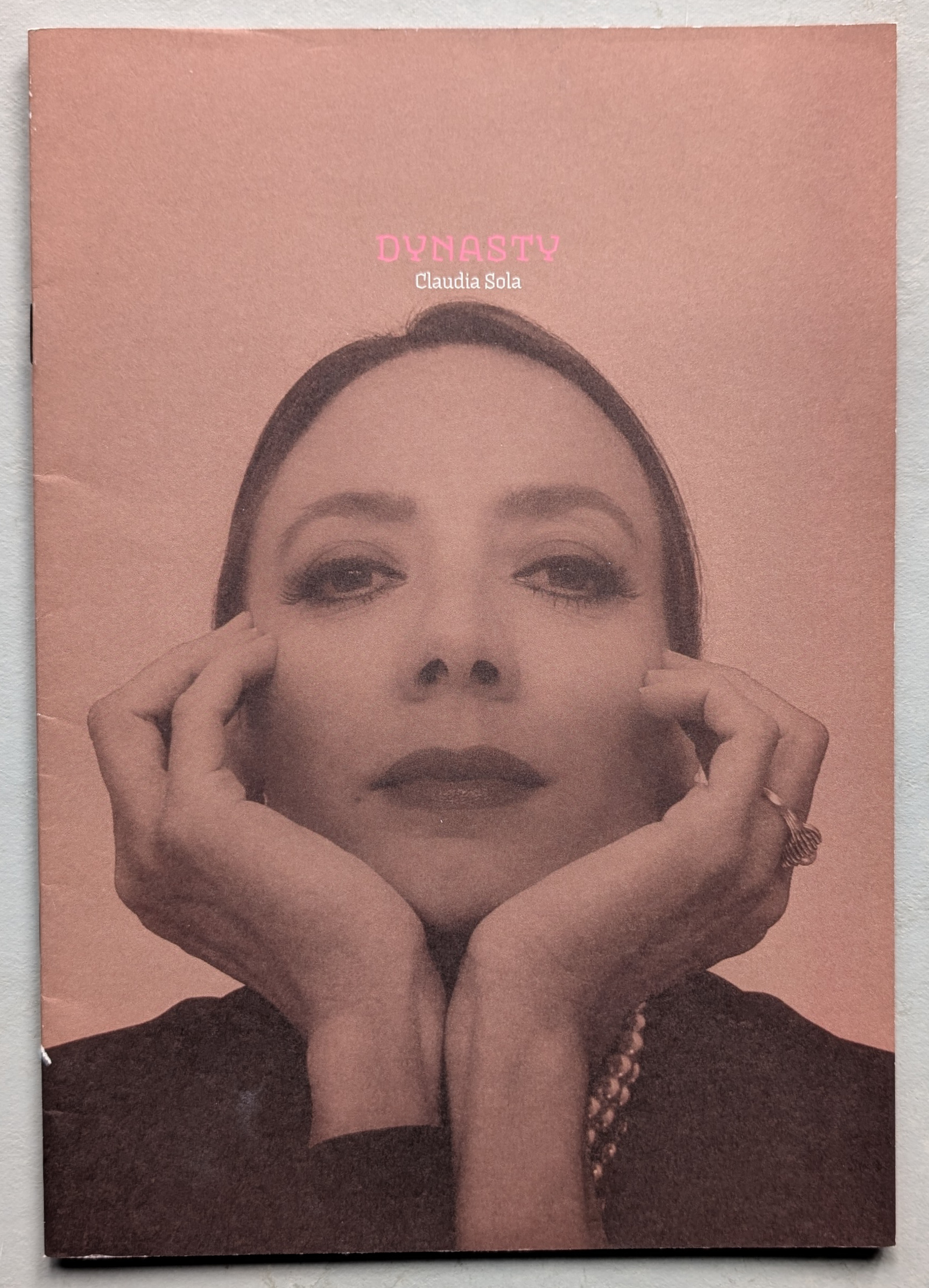 A close-up portrait of Claudia Sola, featuring her hands framing her face and looking directly into the camera. The background is a soft pink color, and the text 'DYNASTY' and 'Claudia Sola' is displayed at the top.