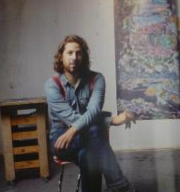 A man with long hair and a casual outfit sitting on a chair in front of a colorful artwork against a wall.