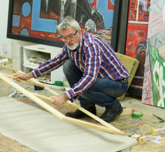 An elderly man with a grey beard and glasses, wearing a red and blue checkered shirt, kneels on the floor of an art studio, manipulating a wooden frame over a canvas.