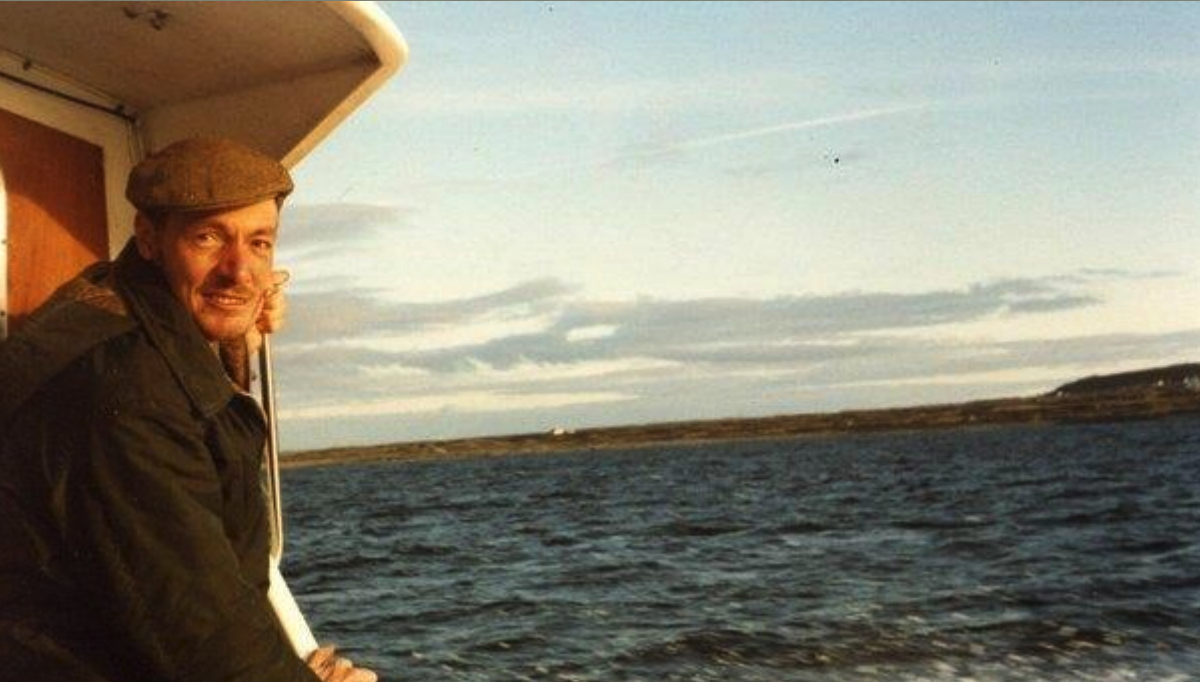 A smiling man wearing a flat cap sits by the railing of a boat, looking out at the water with a scenic view of the horizon and clouds in the background.