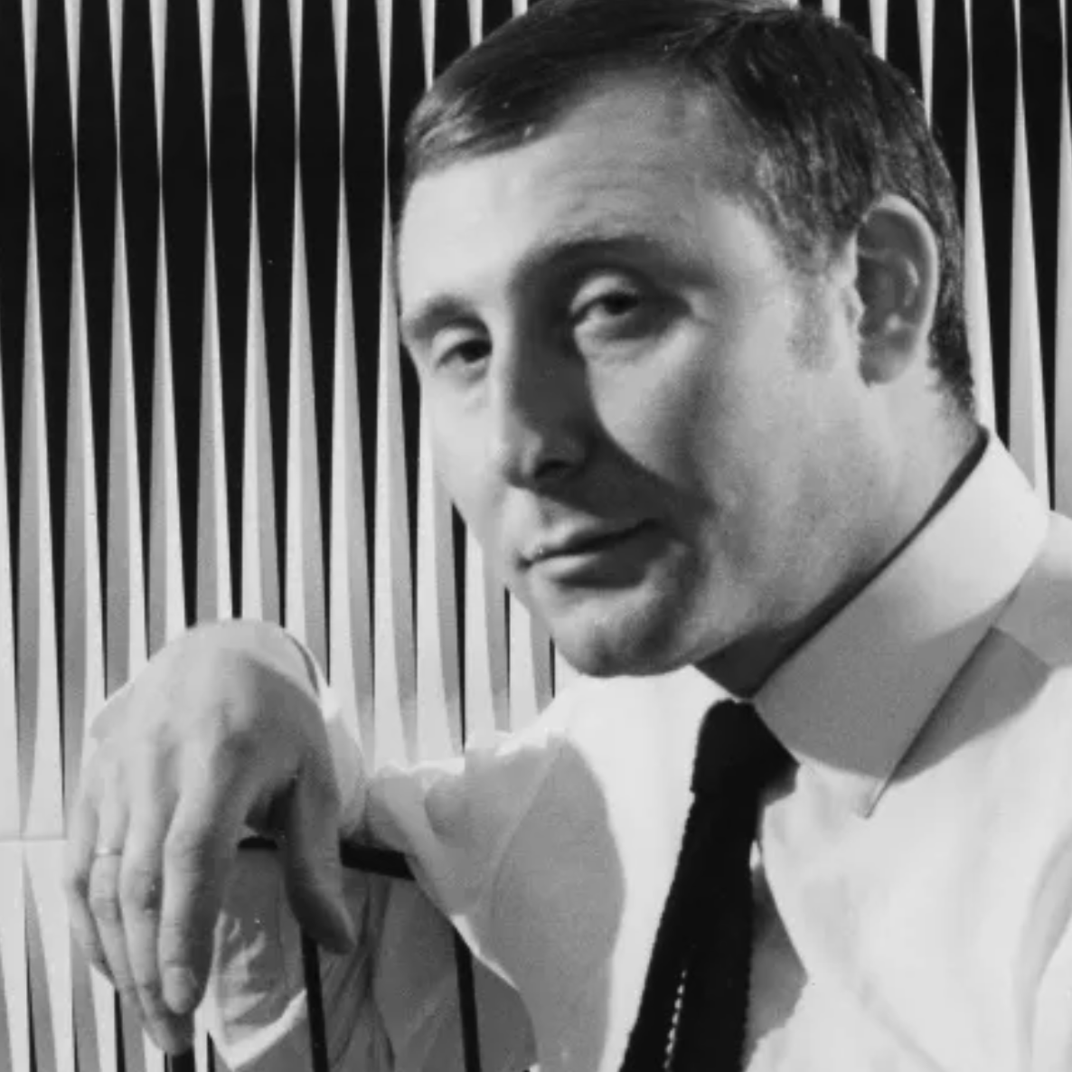 A man in a black and white photo, wearing a white shirt and black tie, leaning against a chair with vertical lines in the background.