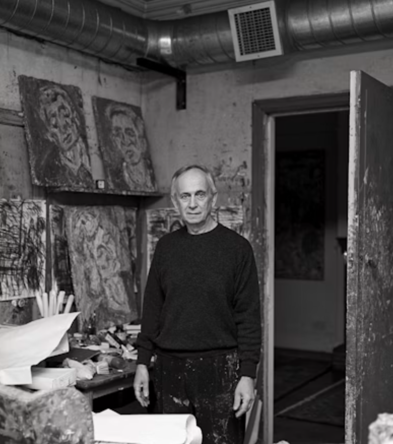 A black and white image of an artist standing in a cluttered studio filled with paintings and art supplies. The artist, wearing a dark sweater and paint-splattered pants, looks directly at the camera.