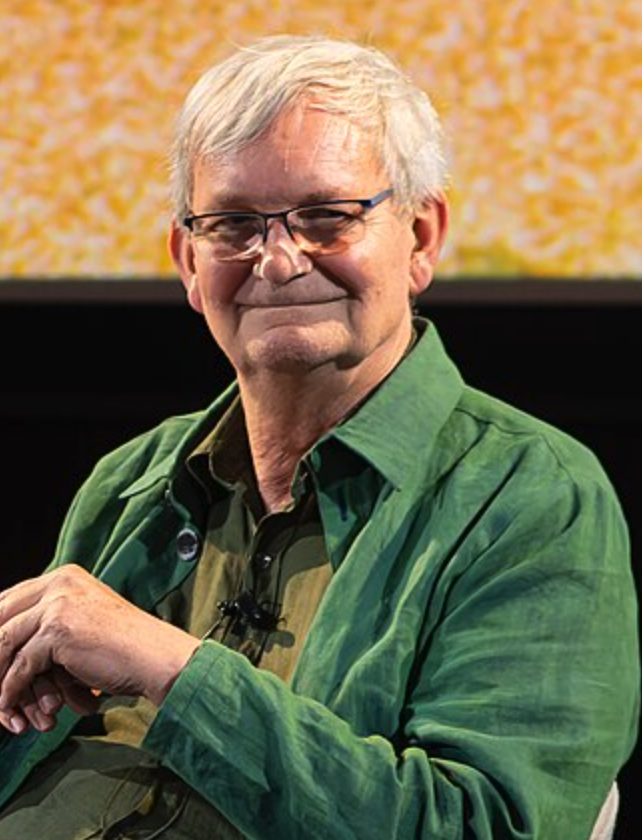A smiling elderly man with glasses, wearing a green jacket, seated against a brightly colored background.
