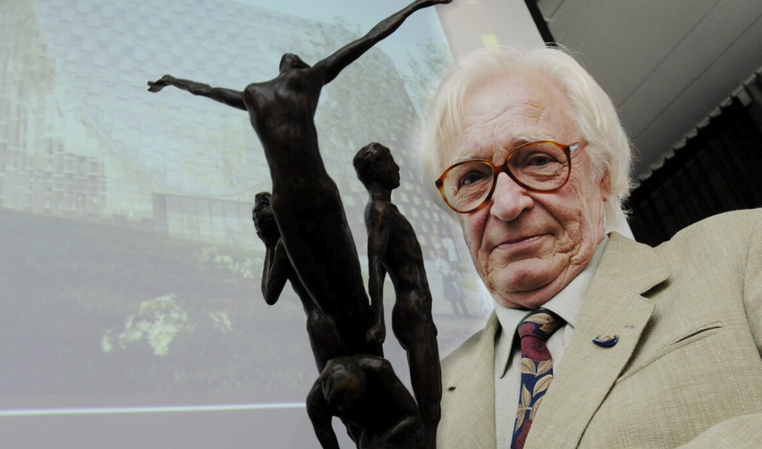 An elderly man with white hair and glasses holds a bronze sculpture depicting three figures, while a building in the background is partially visible.
