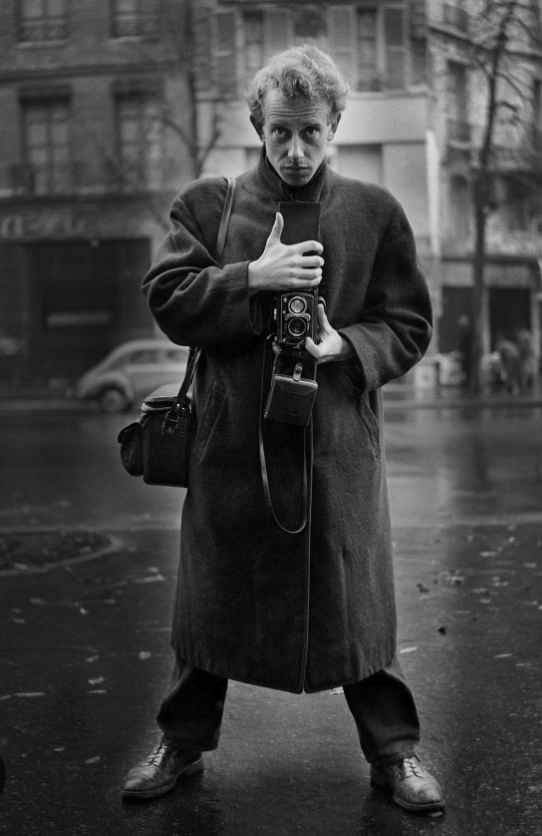 A man wearing a large coat stands on a wet street, holding a camera and looking directly at the viewer.