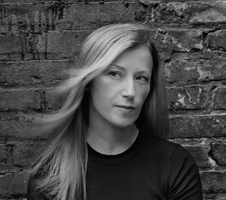 Black and white portrait of a woman with long hair, looking thoughtfully to the side against a textured brick wall.