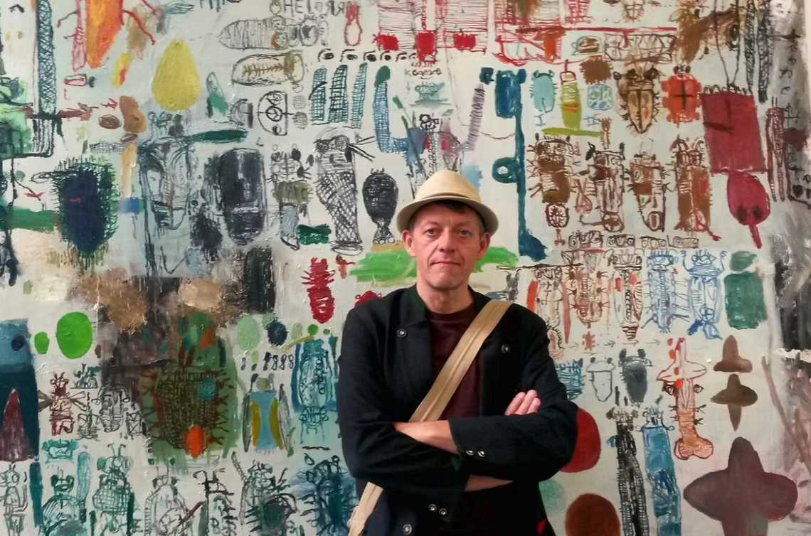 A man in a hat with crossed arms stands in front of a colorful, abstract mural filled with various drawings and designs.