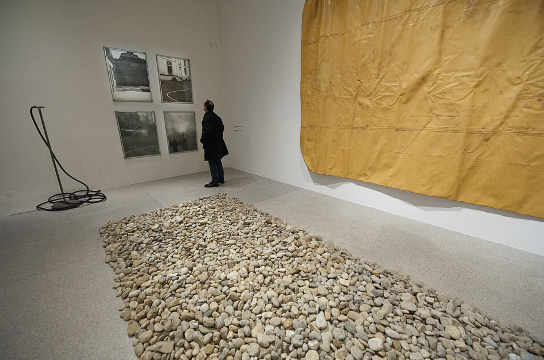 A person observes artwork in an art gallery, featuring black and white photographs on the wall and a large yellow canvas hanging nearby, with a path of smooth stones on the floor.