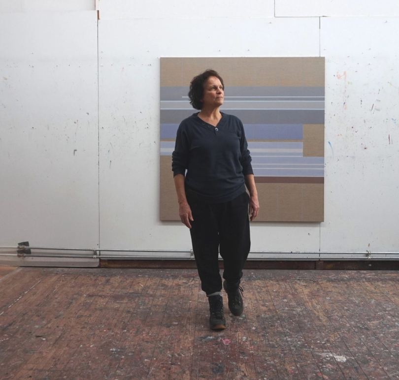 A woman standing in an art studio, wearing a dark top and pants, with a painting featuring horizontal stripes in the background.