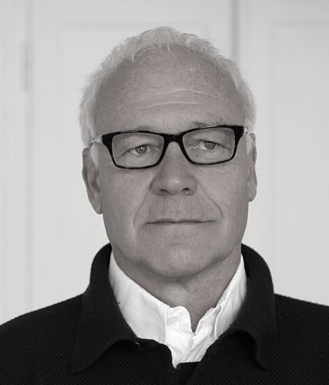 A portrait of a middle-aged man with white hair and glasses, wearing a dark sweater over a white shirt.