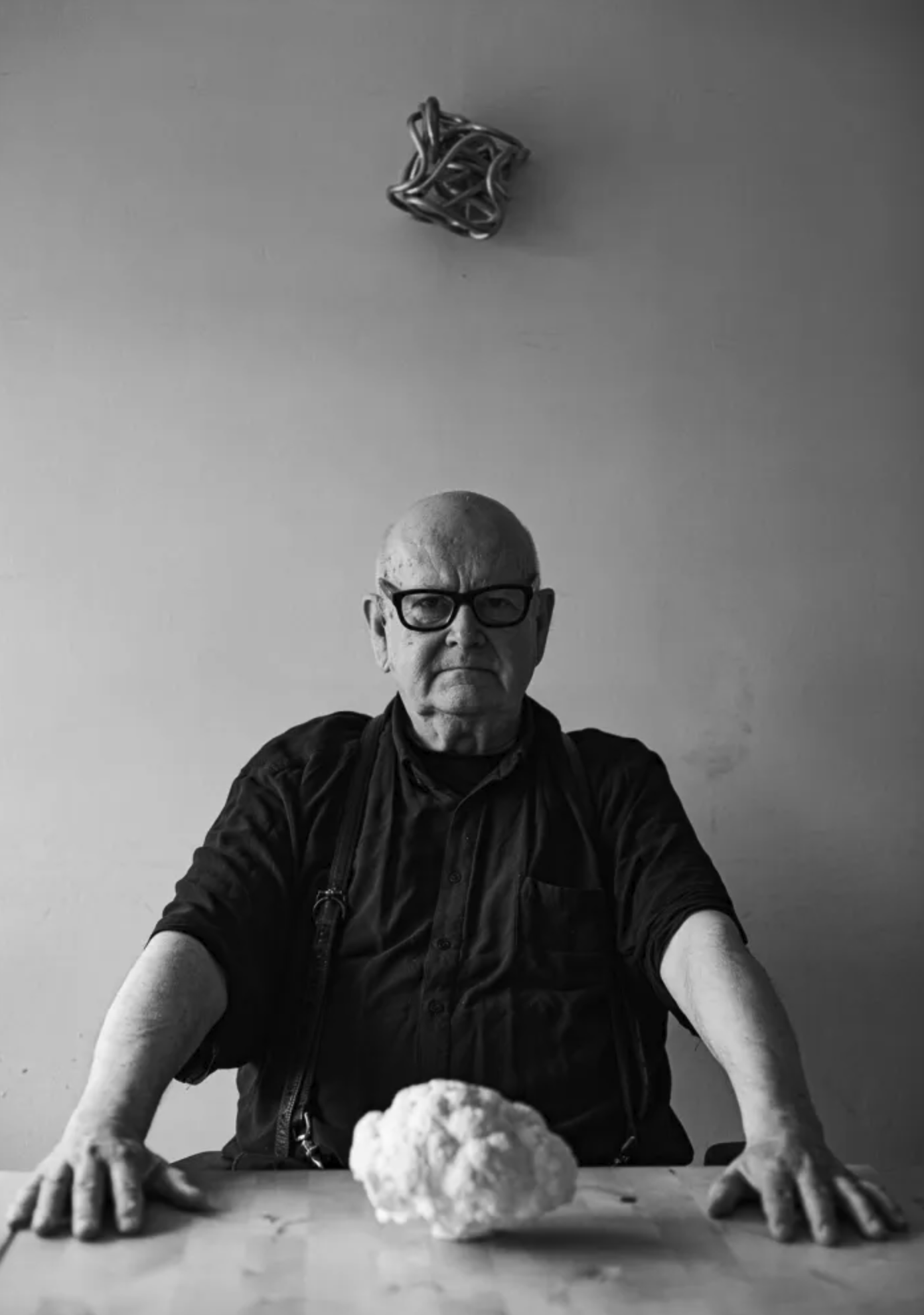 An elderly man wearing glasses sits at a wooden table with his arms extended, flanking a small, white sculptural object. The background is plain and light-colored, creating a stark contrast with the subject.