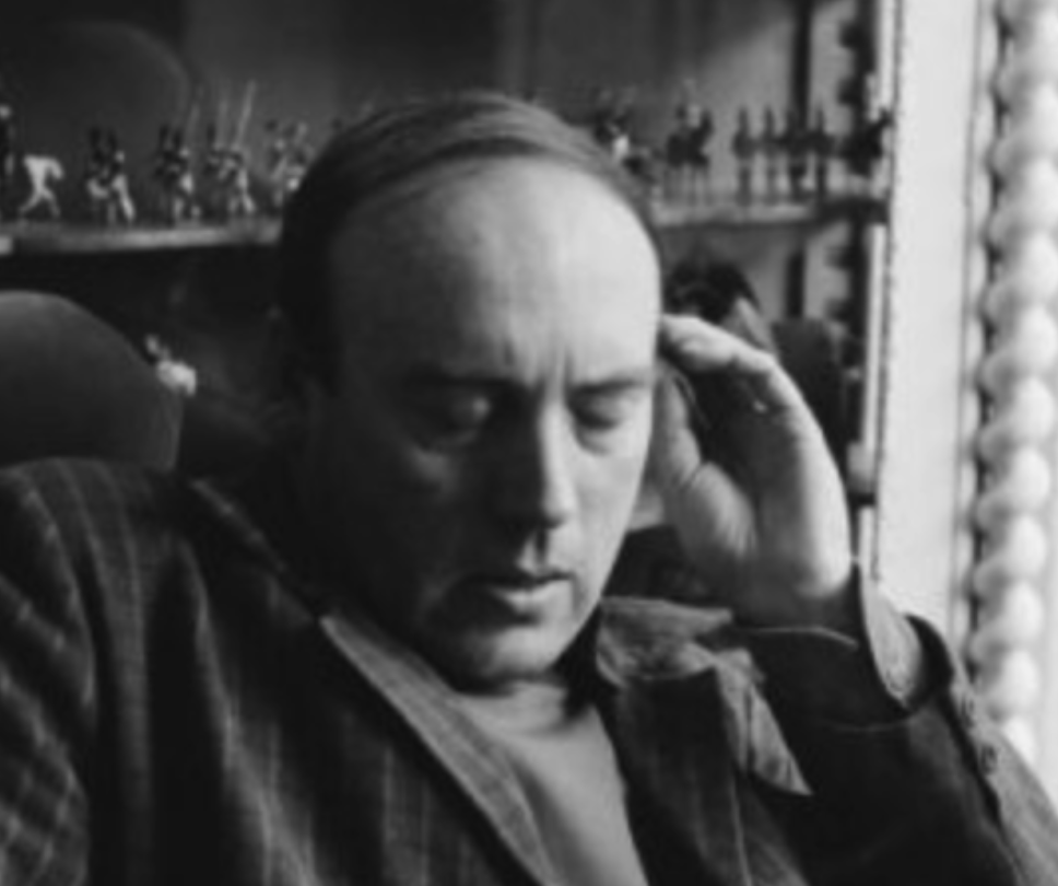 A black and white portrait of a man with closed eyes, deep in thought, wearing a striped jacket, sitting against a backdrop of small figurines.