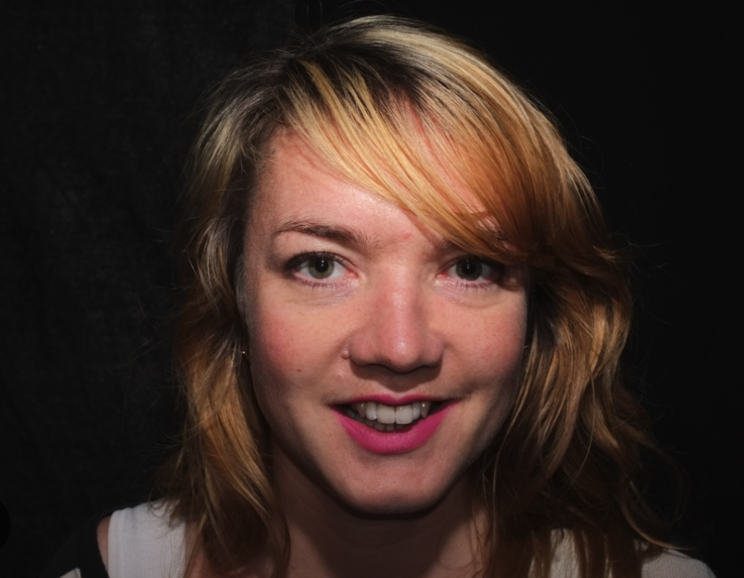 Close-up portrait of a smiling woman with blonde hair and pink lipstick against a dark background.