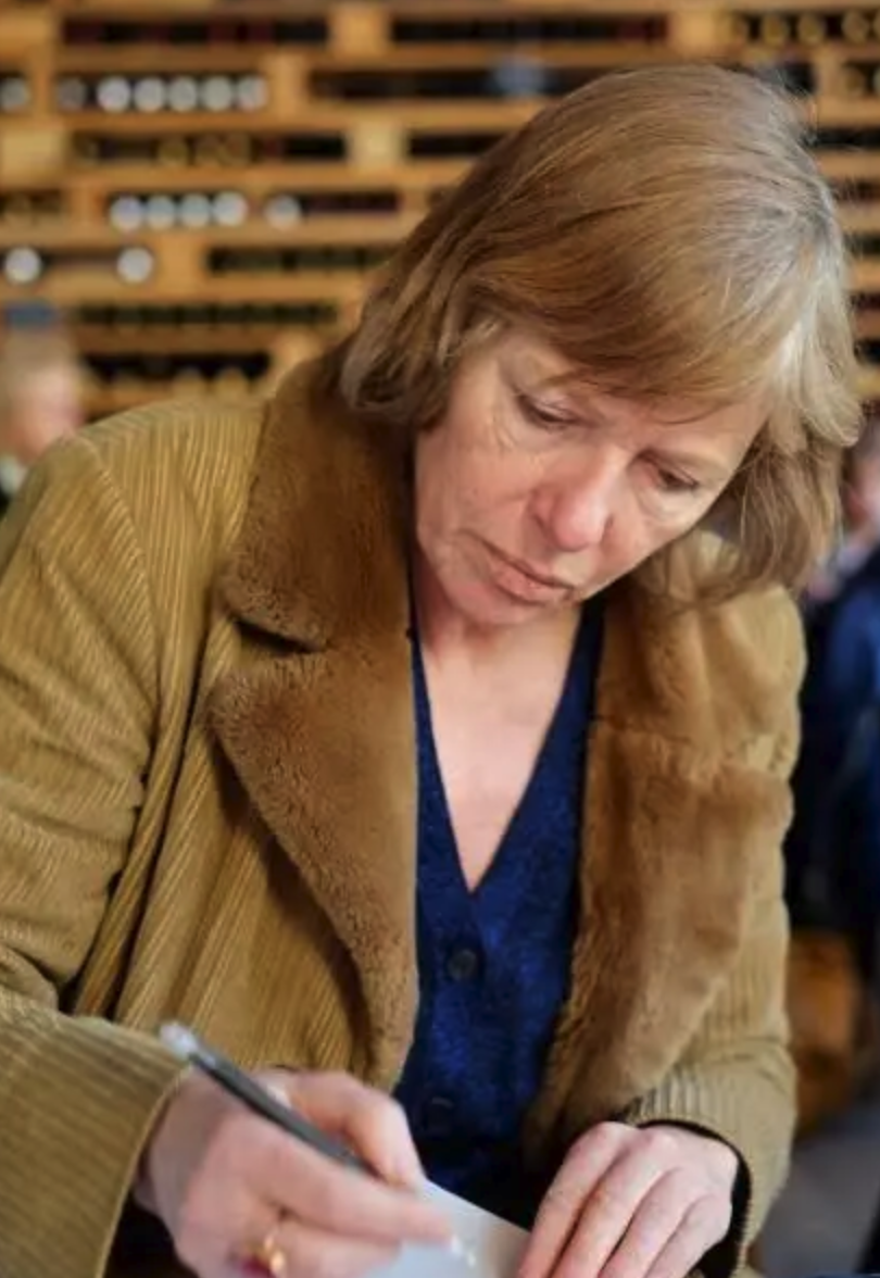 A woman with short brown hair, wearing a corduroy jacket with a fur collar, is focused on writing on a piece of paper.