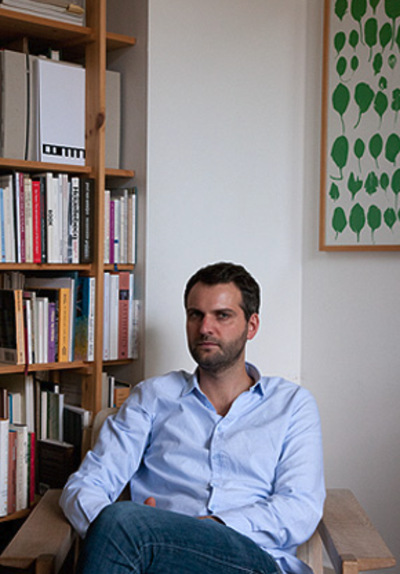A person sitting in a chair, facing the camera, with bookshelves filled with various books in the background and a decorative framed artwork featuring green leaves on the wall.