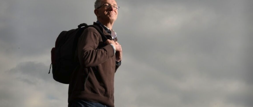 A man standing outdoors with a backpack, looking confidently towards the horizon against a cloudy sky.