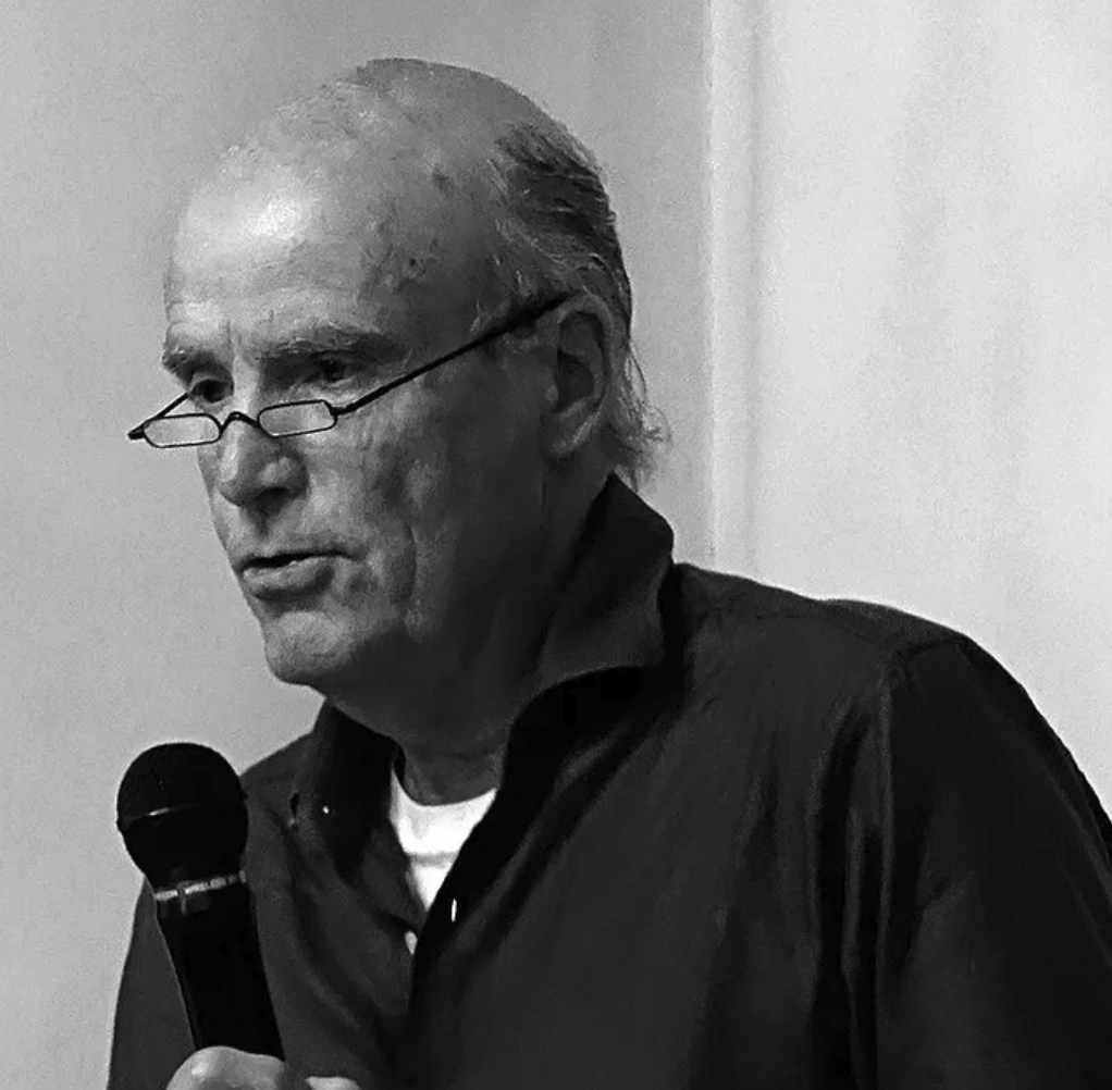 A man with a bald head and glasses speaking into a microphone, wearing a dark shirt.