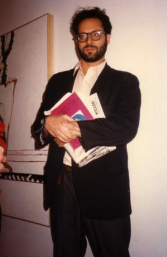 A man in glasses and a suit holds several books and folders, standing in front of an abstract artwork.