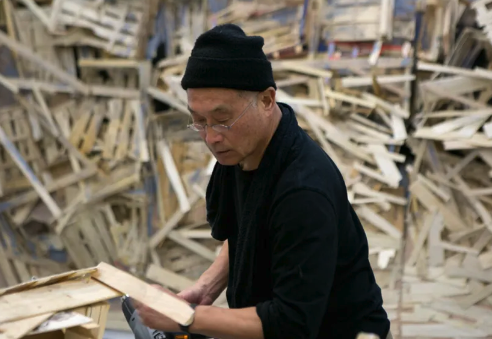 An artist in a black outfit and beanie is focused on assembling wooden pieces in front of a chaotic background made of wooden pallets.