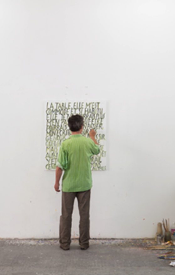 An artist in a green striped shirt is standing in front of a large canvas with text written on it, appearing to paint or add to the artwork. The studio has a minimalistic aesthetic with a clean white background and scattered tools on the floor.