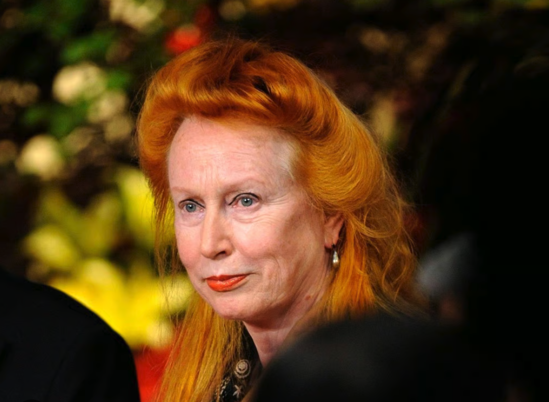 Close-up portrait of a woman with bright orange hair and light makeup, looking thoughtfully at the camera, with a blurred background of plants.