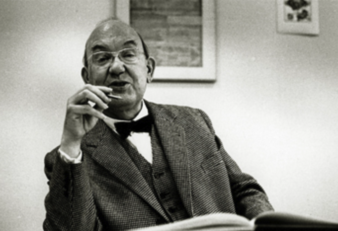 A middle-aged man in a suit with a bow tie gestures while speaking, seated at a table with an open book in front of him.