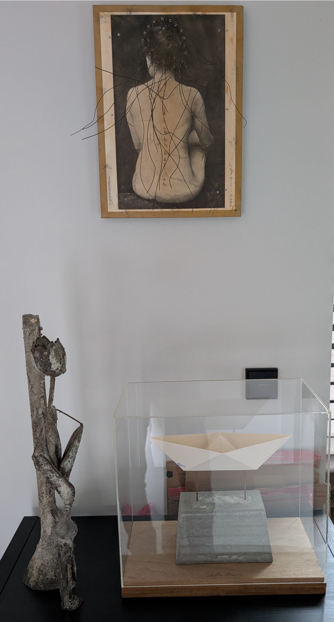 A display showcasing a sculpture, 'Stell Maris' by Sigurdur Gudmundsson, featuring a paper-like structure resting on a concrete base, encased in glass, alongside a bronze sculpture and a framed artwork of a bare back with long hair.