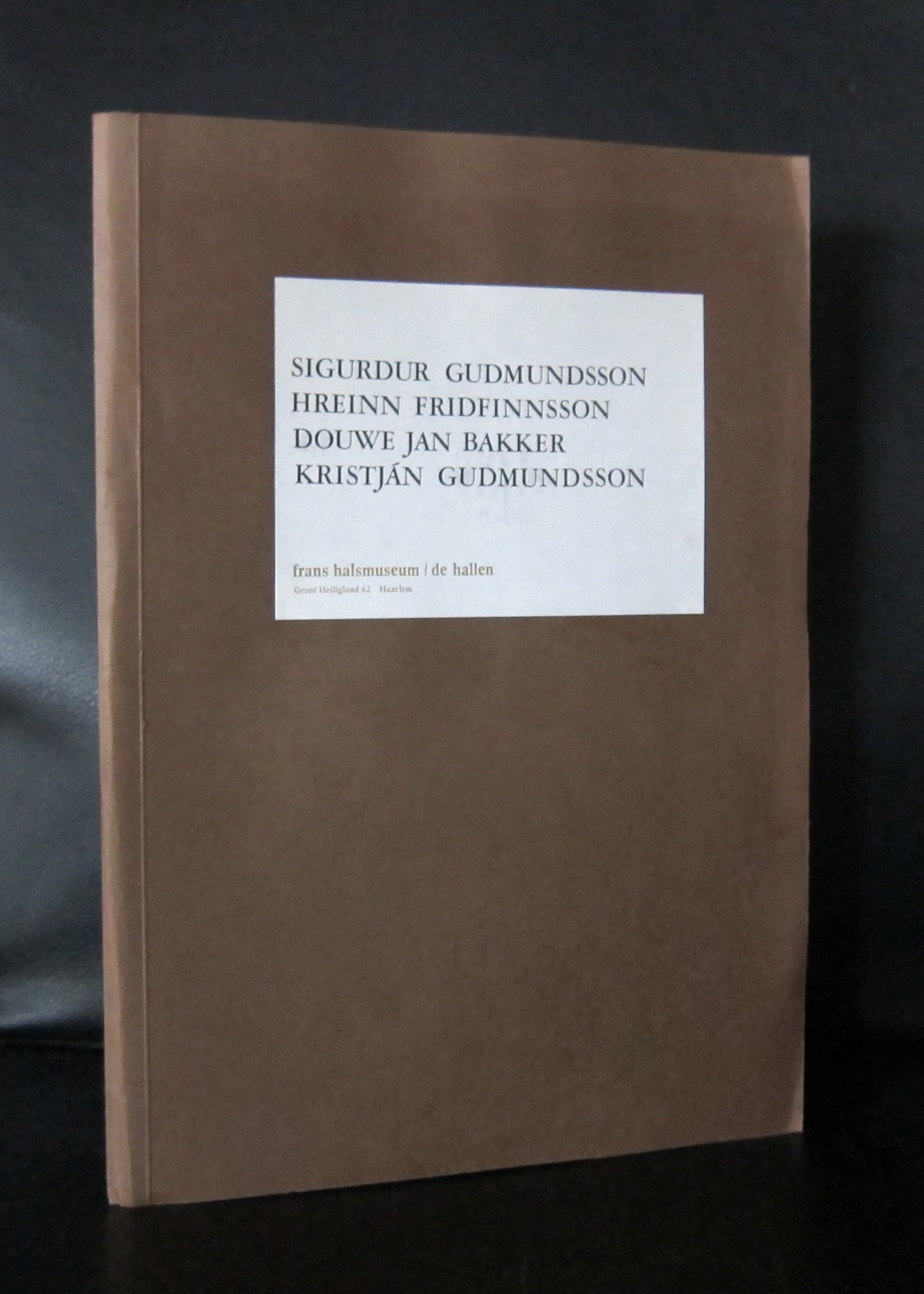 A closed book featuring the title 'Sigurdur Gudmundsson' and the names of other artists, placed on a dark surface against a black background.