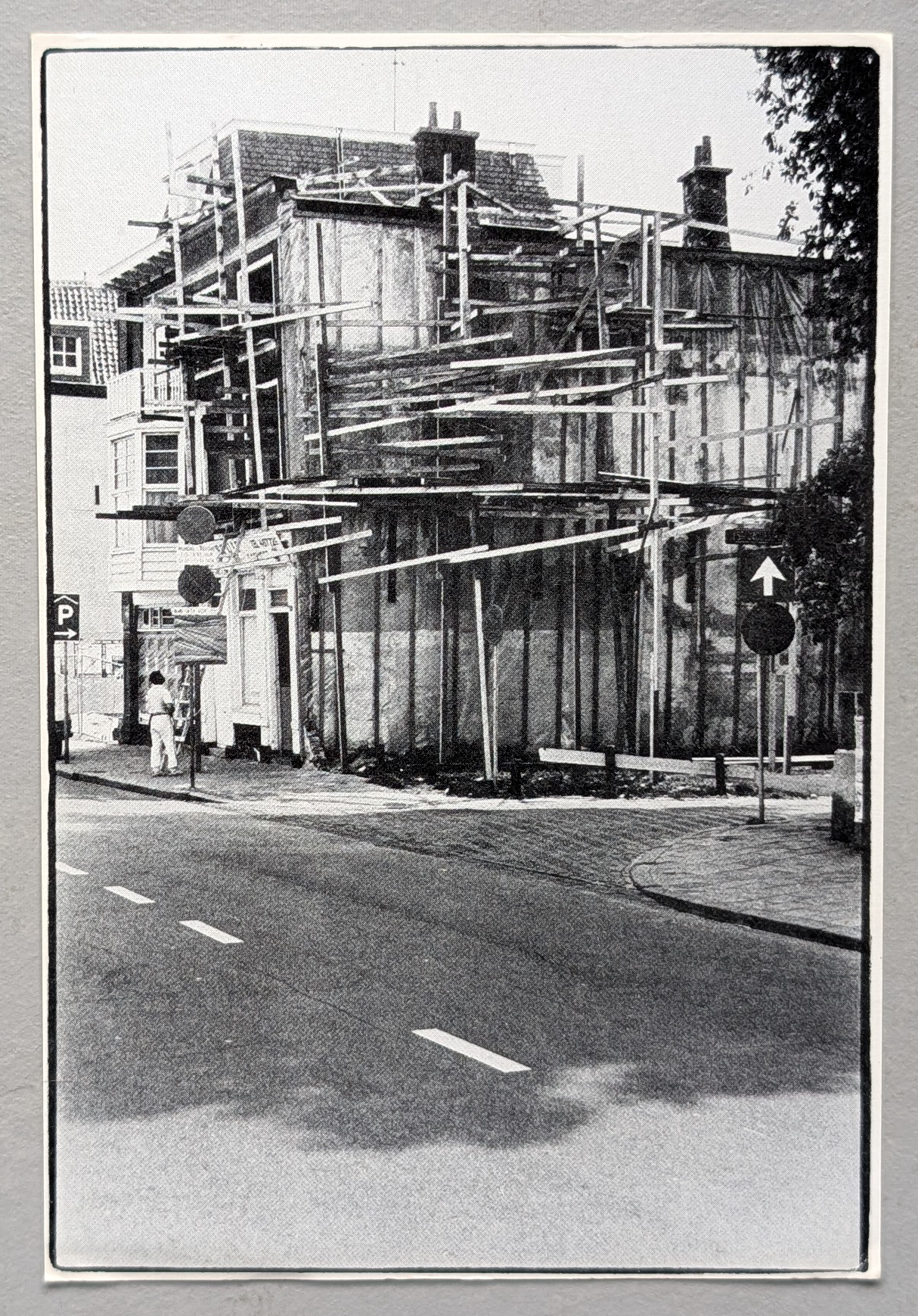 A black and white photograph of a building under construction, surrounded by scaffolding and street signs, with a figure in the foreground observing the site.