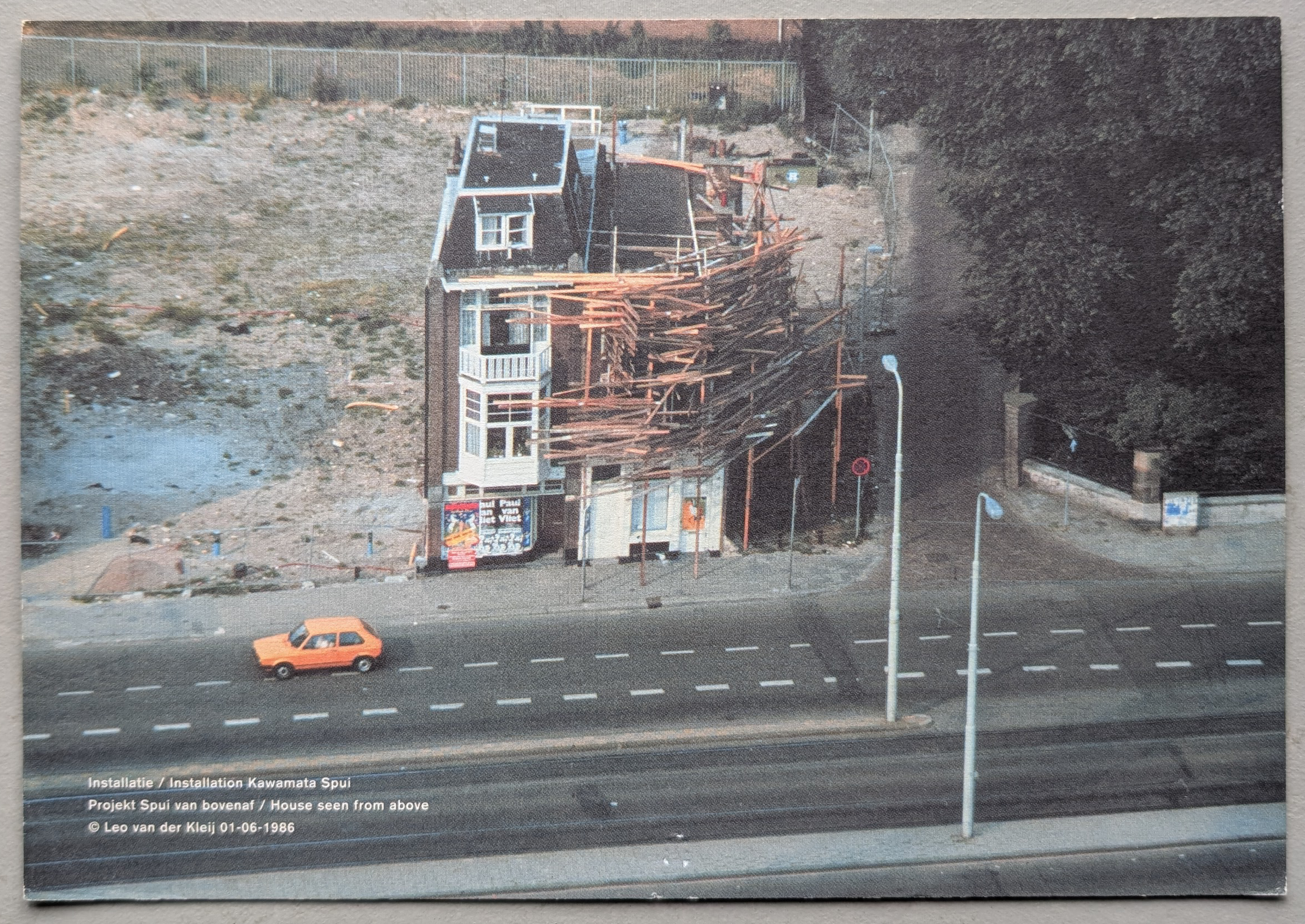 Aerial view of a house installation by Tadashi Kawamata, featuring wooden planks attached to the exterior, with an orange car driving on the street below.