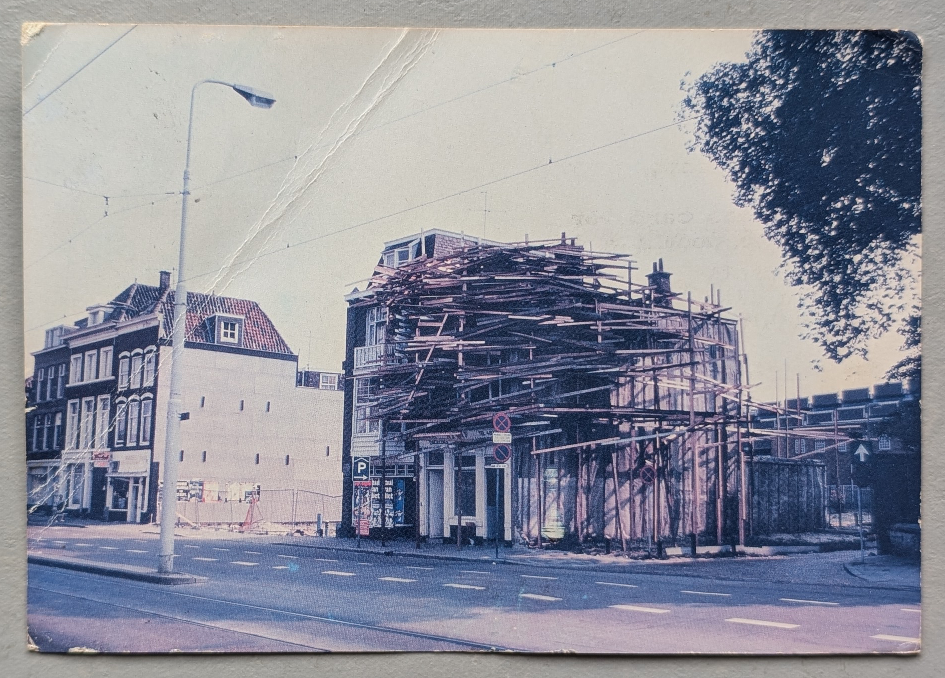 A building with wooden scaffolding artistically arranged around it, set in an urban environment with nearby structures and street elements.