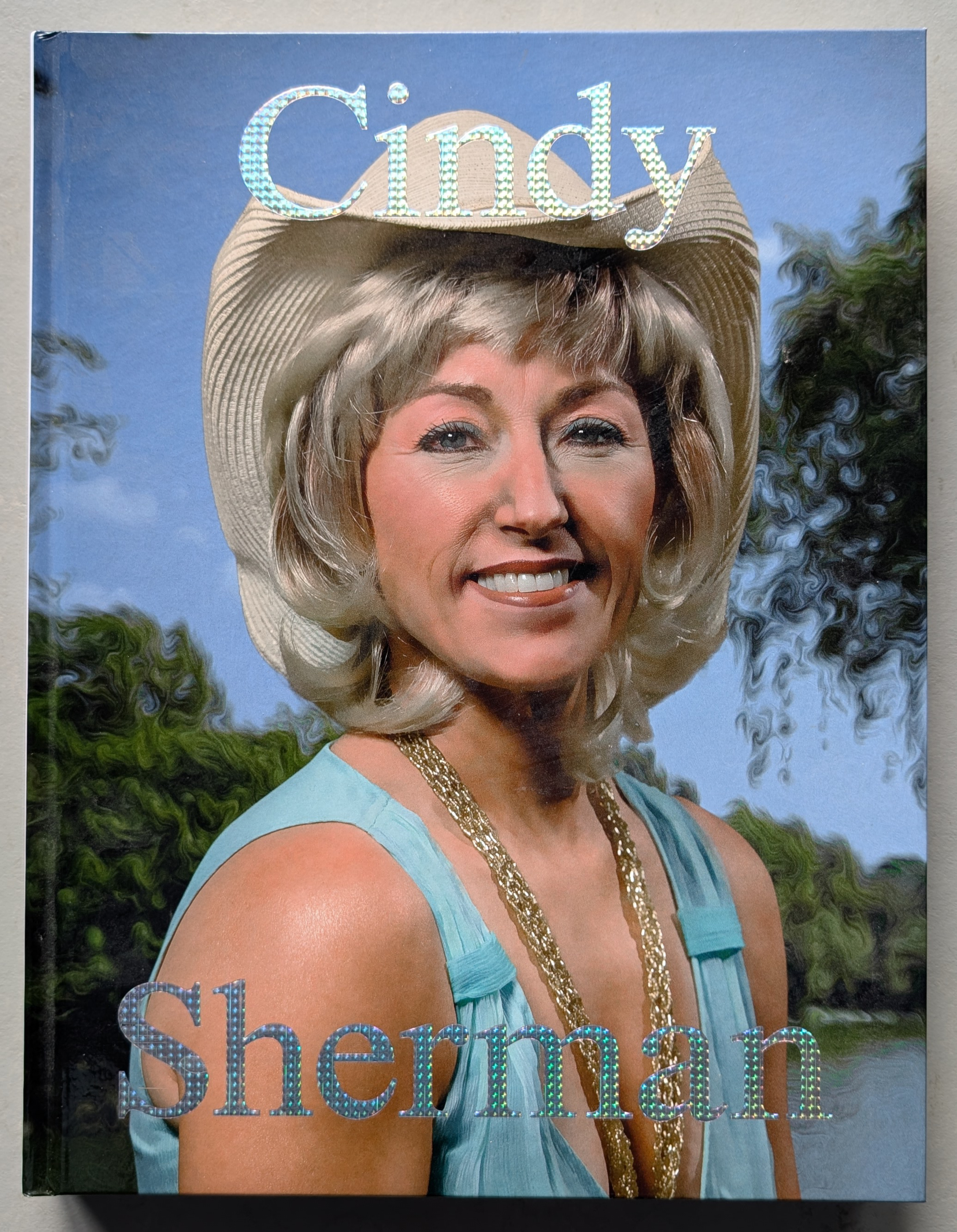 A book cover featuring a smiling woman with blonde hair and a wide-brimmed hat, dressed in a turquoise outfit and adorned with gold jewelry, set against a blurred outdoor background.