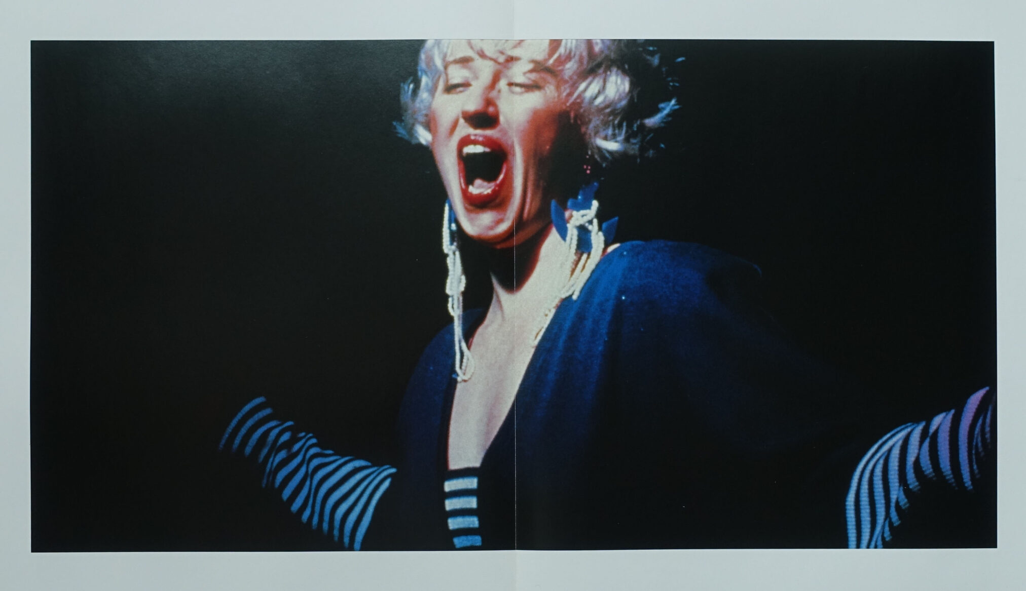 A dramatic, expressive image of a performer with short, curly blonde hair, passionately singing or shouting. The performer wears bold earrings and a dark outfit featuring striped sleeves, set against a contrasting black background.