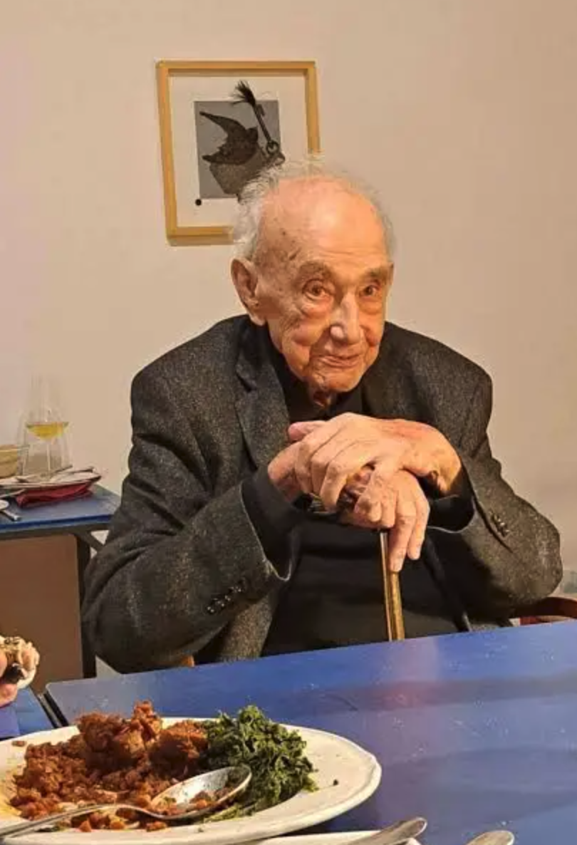 An elderly man with gray hair sits at a table with a cane, looking at the camera. The table has a plate of food, including meat and greens, and a glass is visible in the background.