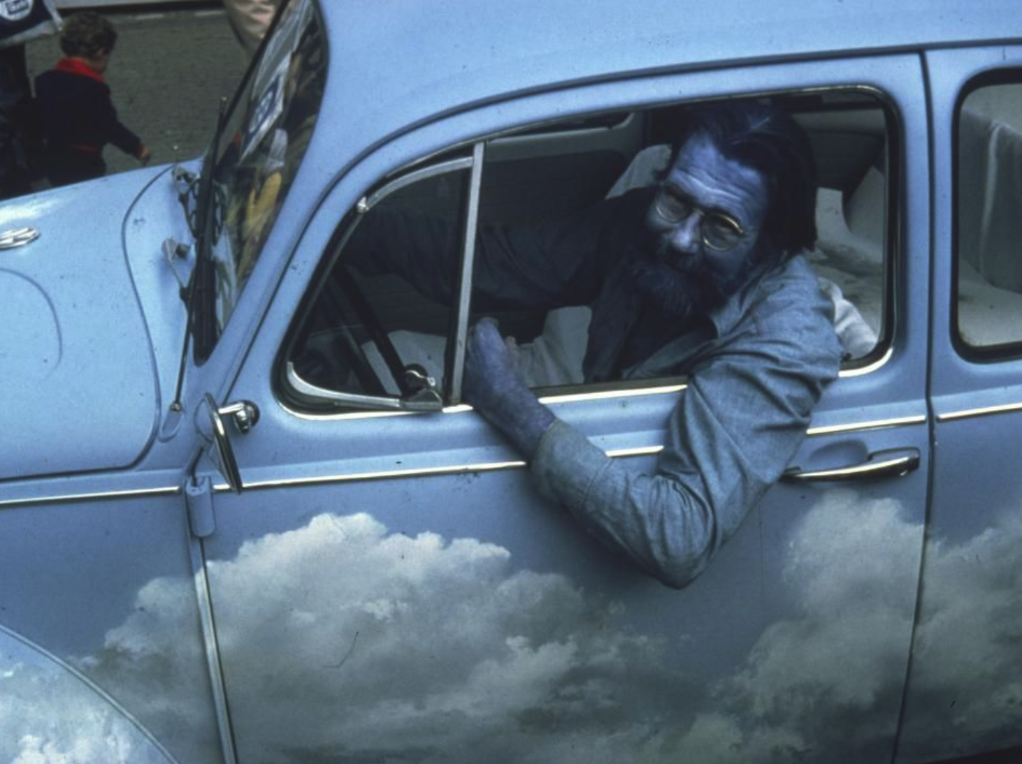 A man with glasses and a beard sitting in a vintage blue car, which has a cloud design on its side.