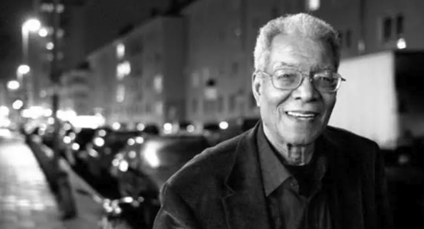 A smiling elderly man wearing glasses and a dark blazer, standing on a city street at night, with blurred cars and buildings in the background.