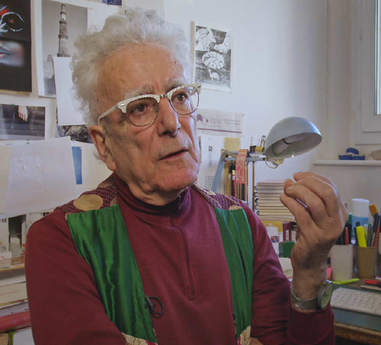 An elderly man with glasses and white hair, dressed in a colorful sweater, gestures while speaking in a studio filled with art supplies and papers.