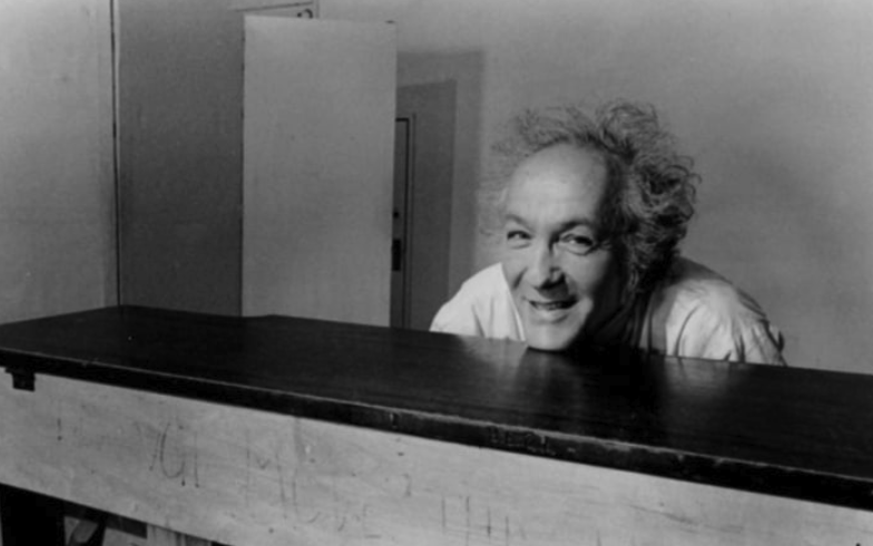 A smiling man with wild hair leaning over a wooden table, captured in black and white.