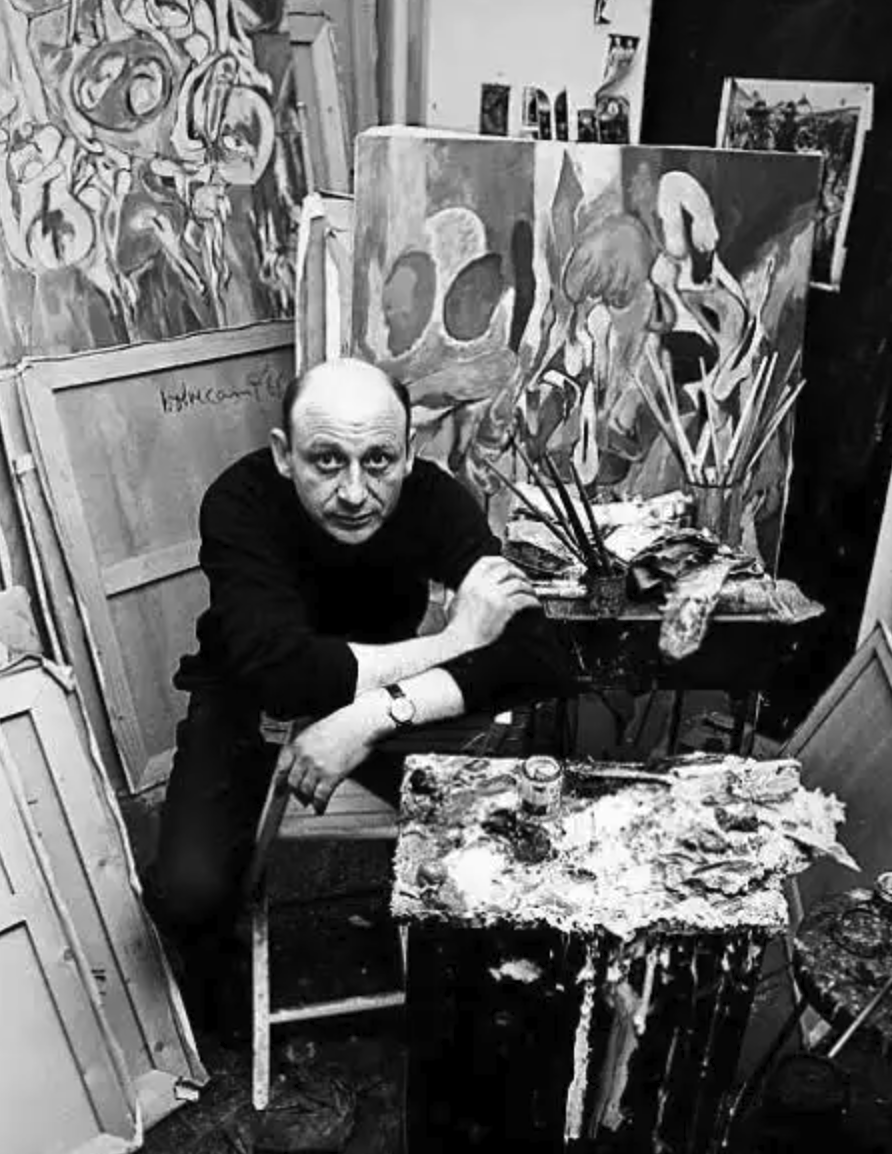 A male artist sitting in his studio surrounded by canvases and painting supplies, with a focus on his expression and work area.