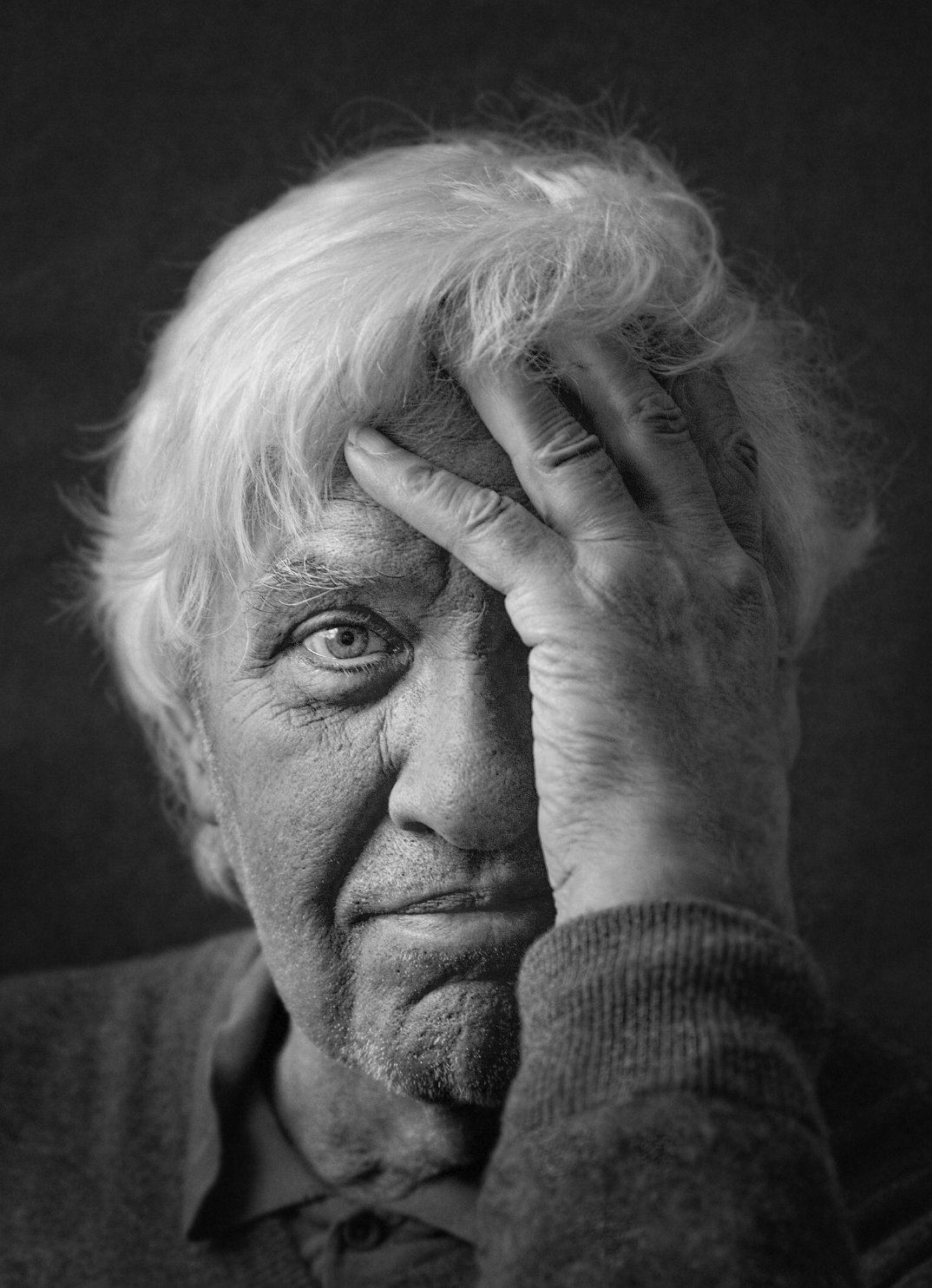 A black and white portrait of an older man with white hair, partially obscured by his hand resting on his forehead, displaying a thoughtful or pensive expression.