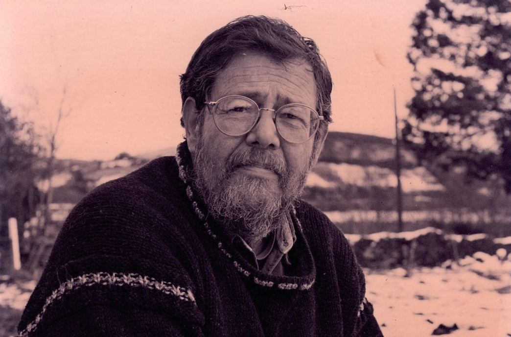 Portrait of an older man with glasses and a beard, wearing a dark sweater, outdoors with a blurred background of snow and trees.