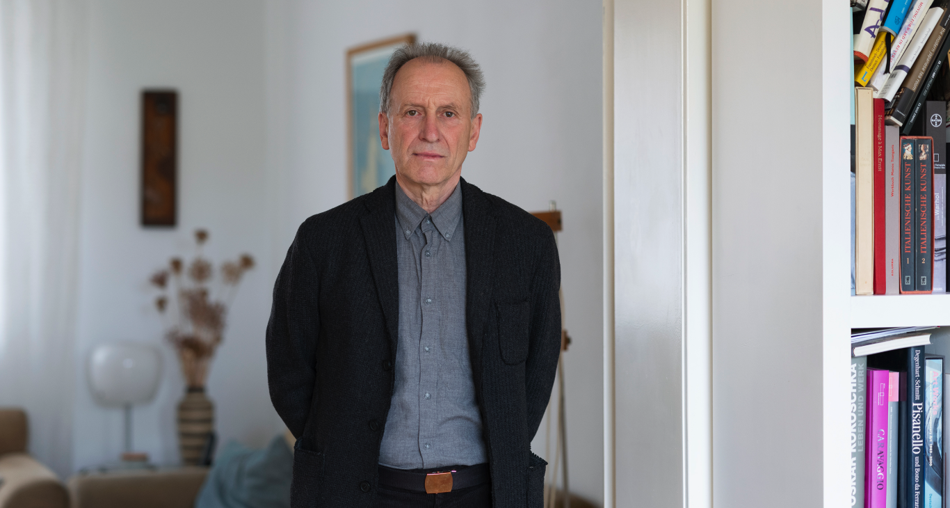 A man standing in a well-lit room, wearing a dark coat over a light shirt, with a bookshelf filled with books in the background.